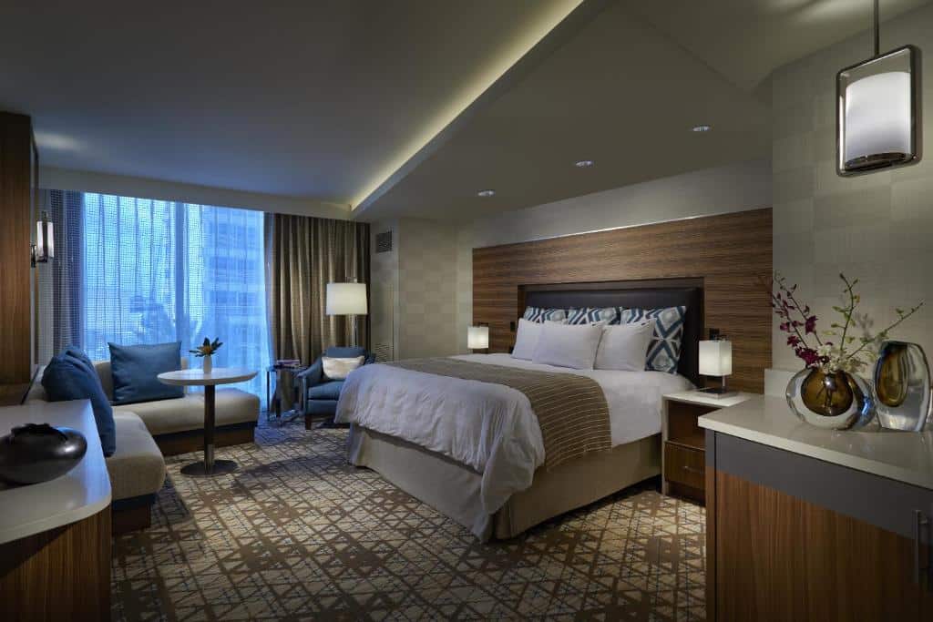 Quarto amplo no Seminole Hard Rock Hotel and Casino Tampa com uma cama de casal, um poltrona, um pequeno sofá em L perto da sacada, duas mesinhas de cabeceira com abajures, tudo decorado em tons de marrom claro, bege e branco.