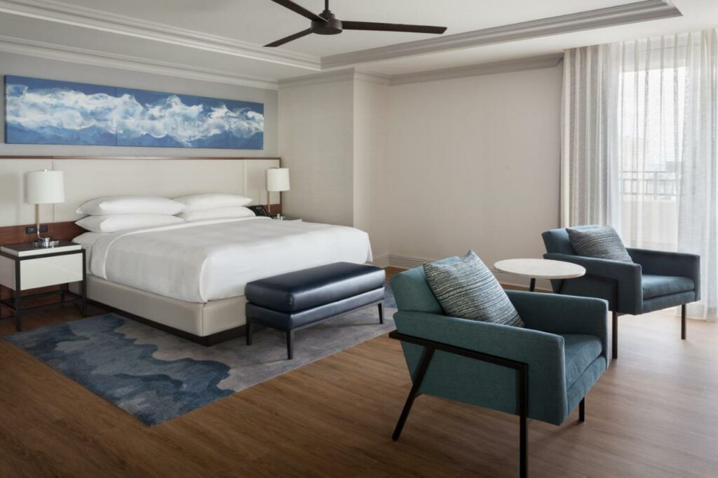 Quarto amplo no Tampa Marriott Water Street com uma cama de casal, duas poltronas, chão de madeira, duas mesinhas de cabeceira com abajures, tudo em tons de azul e verde claro, madeira e branco.