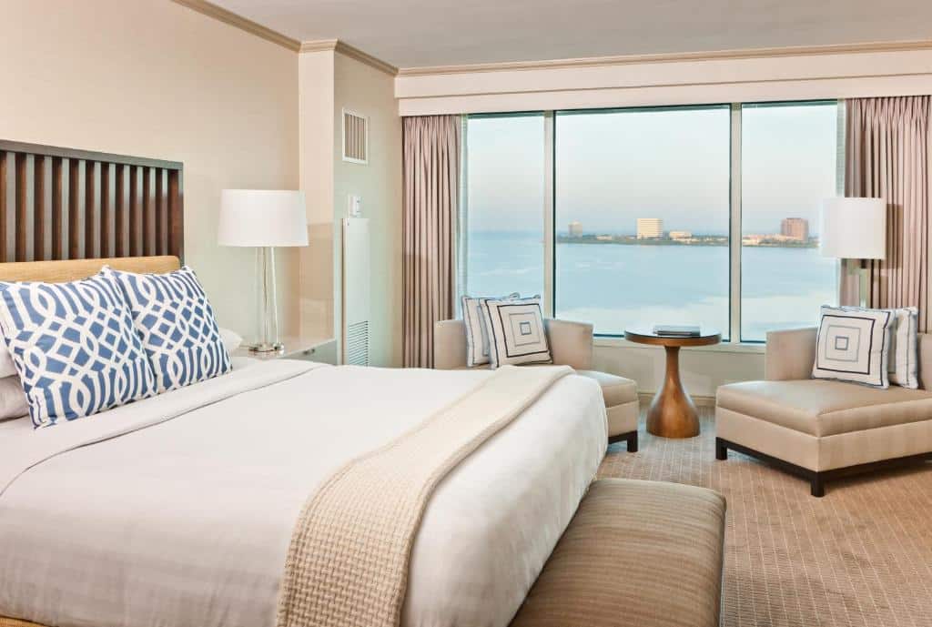 Quarto do Grand Hyatt Tampa Bay com uma cama de casal, uma mesinha com duas poltronas perto de uma janela ampla com vista para o mar, chão de carpete bege, toda a decoração está em tons de bege claro e branco.