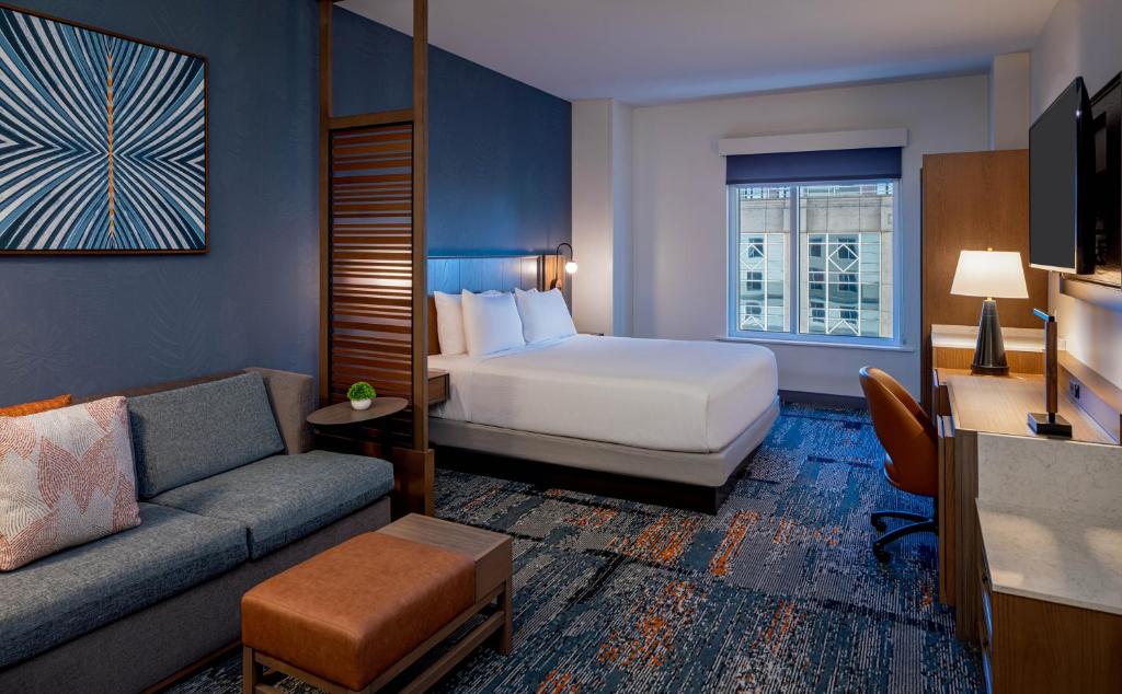 Quarto no Hyatt Place Tampa Downtown com uma cama de casal, uma mesinha de escritório com uma cadeira giratória, uma sofá, uma janela e uma televisão, tudo decorado em tons de marrom, laranja e azul.