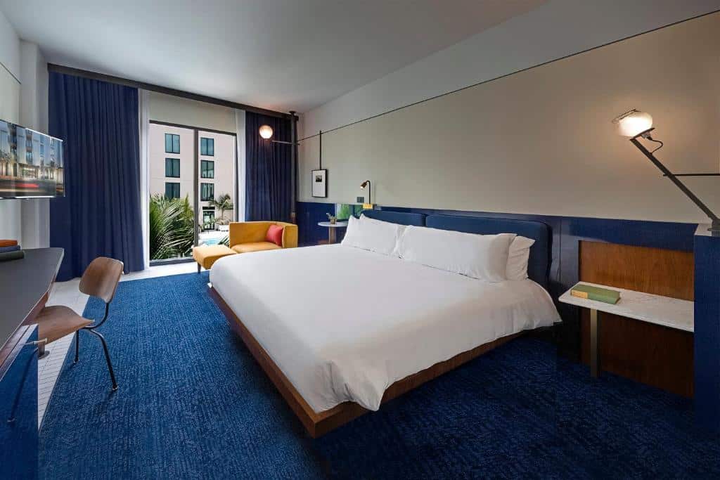 Quarto no Hotel Haya com uma cama de casal, uma poltrona, uma varanda, uma televisão, uma mesa de escritório com uma cadeira, há um carpte azul no local que combina com o restante da decoração que segue o mesmo tom, mesclando com branco
