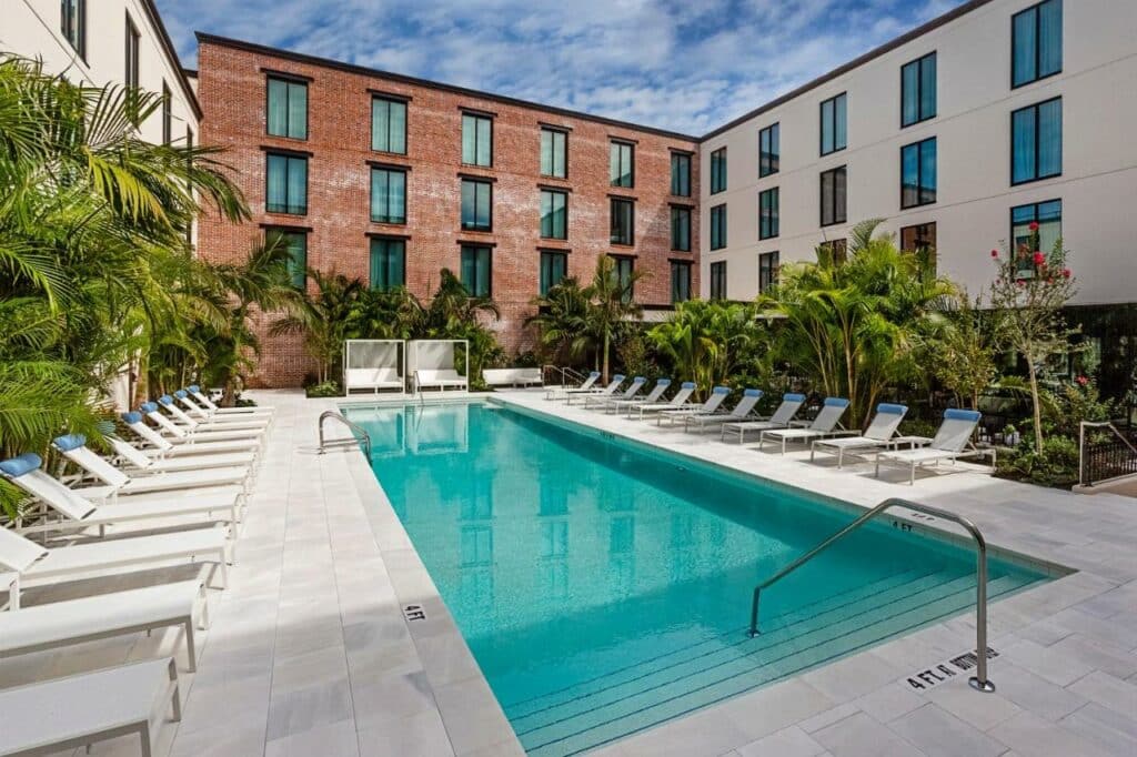 Piscina externa retangular com espreguiçadeiras ao redor, cercada por palmeiras e um prédio moderno de tijolos e branco, na propriedade do Hotel Haya.