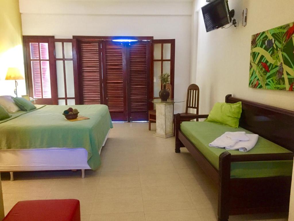 Um quarto com uma cama, um sofá-cama verde, uma cadeira de madeira, uma TV na parede e portas de madeira com persianas na Pousada Caminho do Sol.
