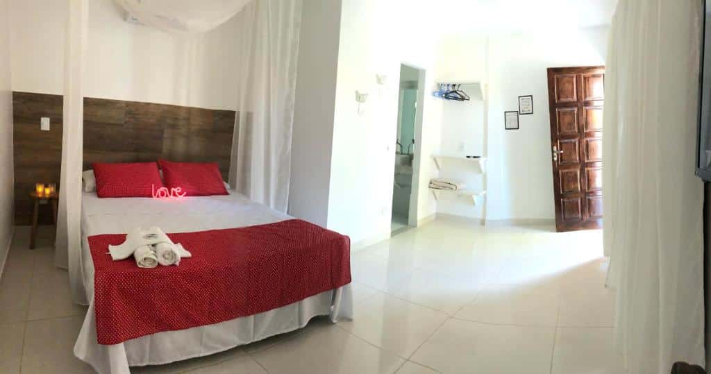 Uma cama bem arrumada com roupa de cama vermelha em um quarto branco e claro com uma porta de madeira e algumas toalhas sobre a cama, no Solar da Praia Pousada.