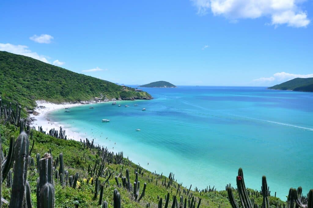 Baía azul-turquesa com barcos, praia de areia, colinas verdes e cactos em primeiro plano sob um céu azul claro em Arraial do Cabo.