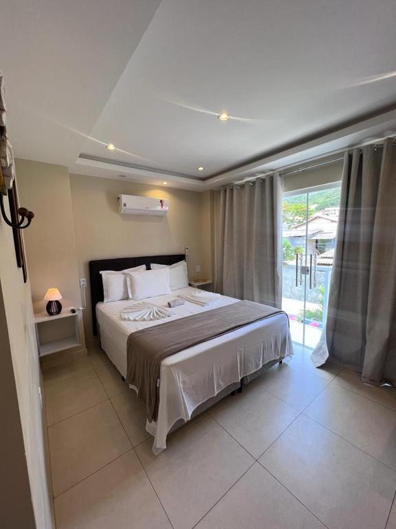 Um quarto na Pousada Arraial Tropical bem arrumado com uma cama de casal, mesinhas de cabeceira e uma grande janela com cortinas cinza, deixando entrar a luz natural.