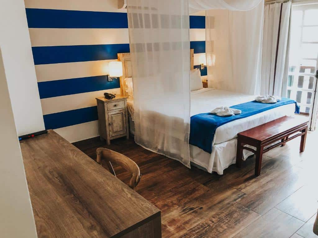 Um quarto na Pousada da Prainha com uma cama de casal, parede listrada de azul, escrivaninha, cadeira e piso de madeira, iluminado por luz natural, para representar pousadas em Arraial do Cabo.