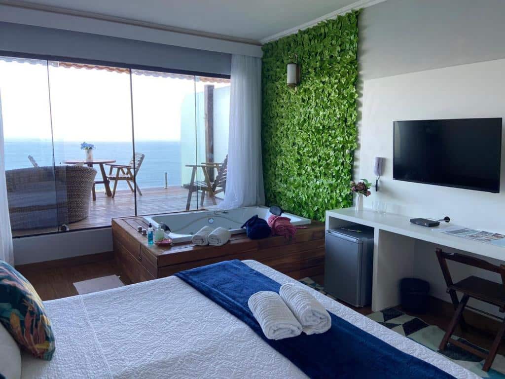 Quarto da Pousada Vista Turquesa com cama, jacuzzi, parede de plantas verdes, TV, frigobar e varanda com vista para o mar, para representar pousadas em Arraial do Cabo.