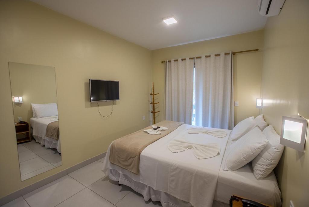Um pequeno quarto de hotel com uma cama de casal, TV na parede, espelho, ar-condicionado e cortinas em uma janela, na Costa Dourada Pousada em Arraial do Cabo.