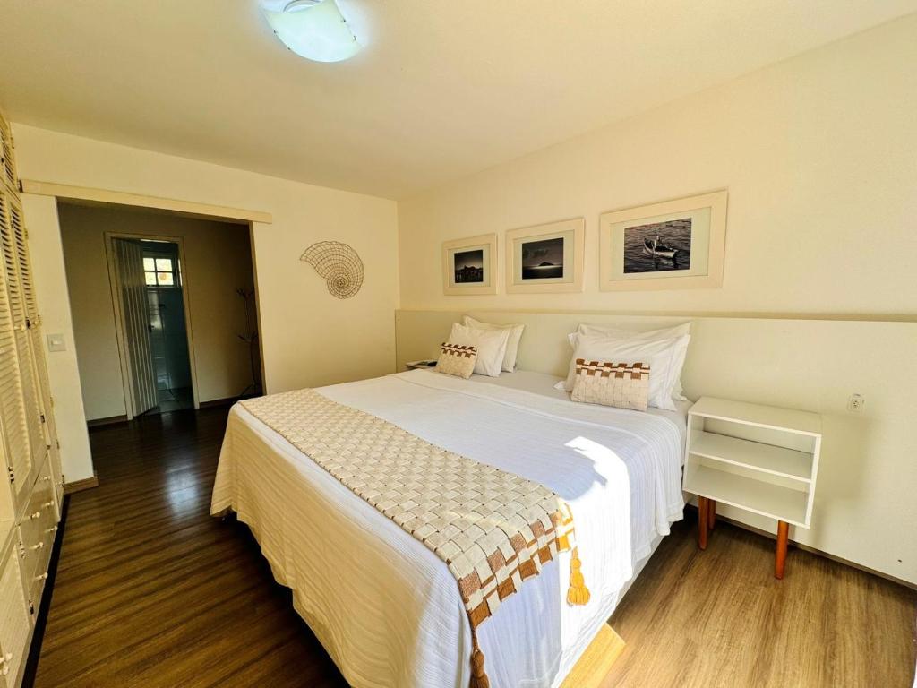 Um quarto com uma cama grande, duas mesinhas de cabeceira brancas, piso de madeira e três fotos emolduradas em uma parede branca na Pousada Arraial Inn, para representar pousadas em Arraial do Cabo.