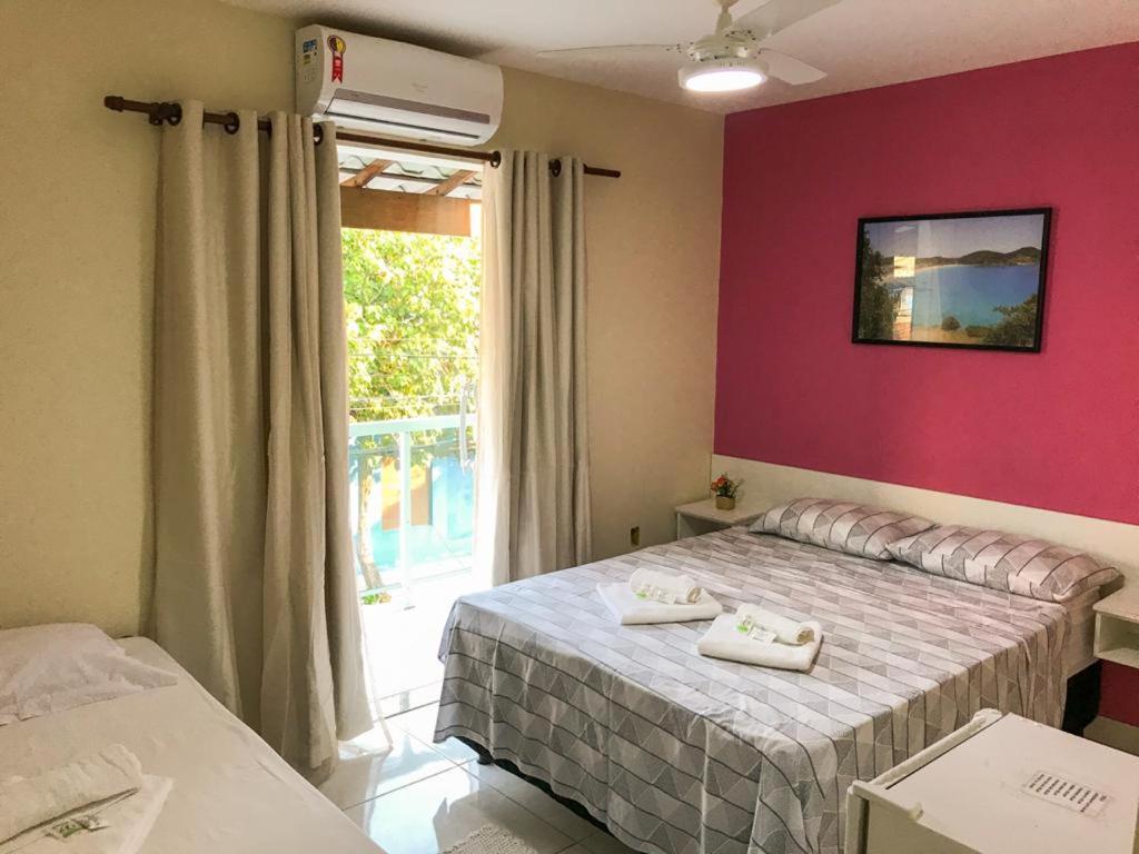 Um quarto bem iluminado com uma cama de casal, uma cama de solteiro, uma porta para a varanda, uma parede com detalhes em rosa e um ventilador de teto, na Pousada Caminho dos Anjos, para representar pousadas em Arraial do Cabo.