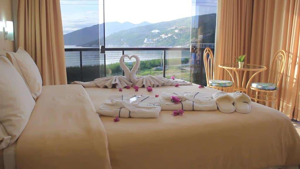 Um quarto na Pousada Canto da Baleia com uma cama de casal, roupões, chinelos, uma pequena mesa com cadeiras e vista para as montanhas e a água, para representar pousadas em Arraial do Cabo.