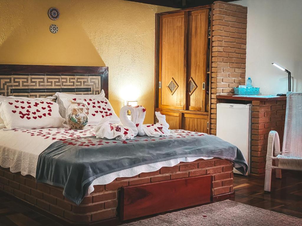 Uma cama bem arrumada com cisnes de toalha e pétalas de rosa, ao lado de uma pequena geladeira, um guarda-roupa e uma lâmpada acesa em uma mesa de cabeceira, na Pousada Espelho Dagua em Gonçalves.