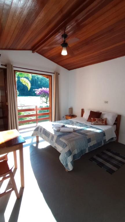 Quarto iluminado pelo sol com teto de madeira, cama de casal, mesa pequena, porta aberta para um jardim e um tapete no chão, na Pousada Vitória, para representar pousadas em Gonçalves.