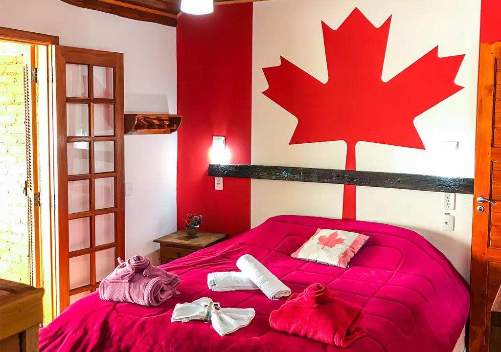 Um quarto com uma colcha vermelha, toalhas na cama e uma grande folha de bordo vermelha pintada na parede atrás da cama, na Pousada Cabanas no Mundo, para representar pousadas em Gonçalves.