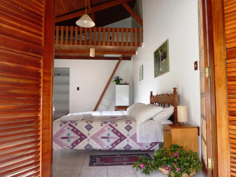 Um quarto com cama de madeira, colcha estampada, loft, teto de madeira e plantas no piso de azulejos, na Pousada Passaredo, para representar pousadas em Gonçalves.