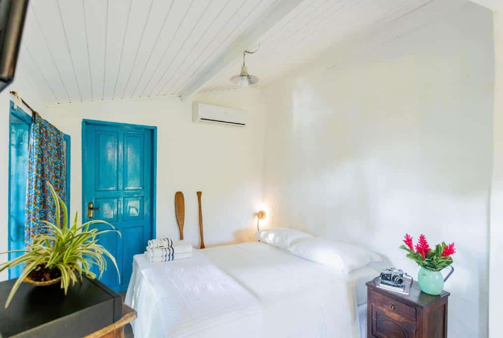 Um quarto branco da pousada Ilhabela Homestay com uma cama de casal, porta azul, móveis de madeira, ar-condicionado e vasos de plantas.