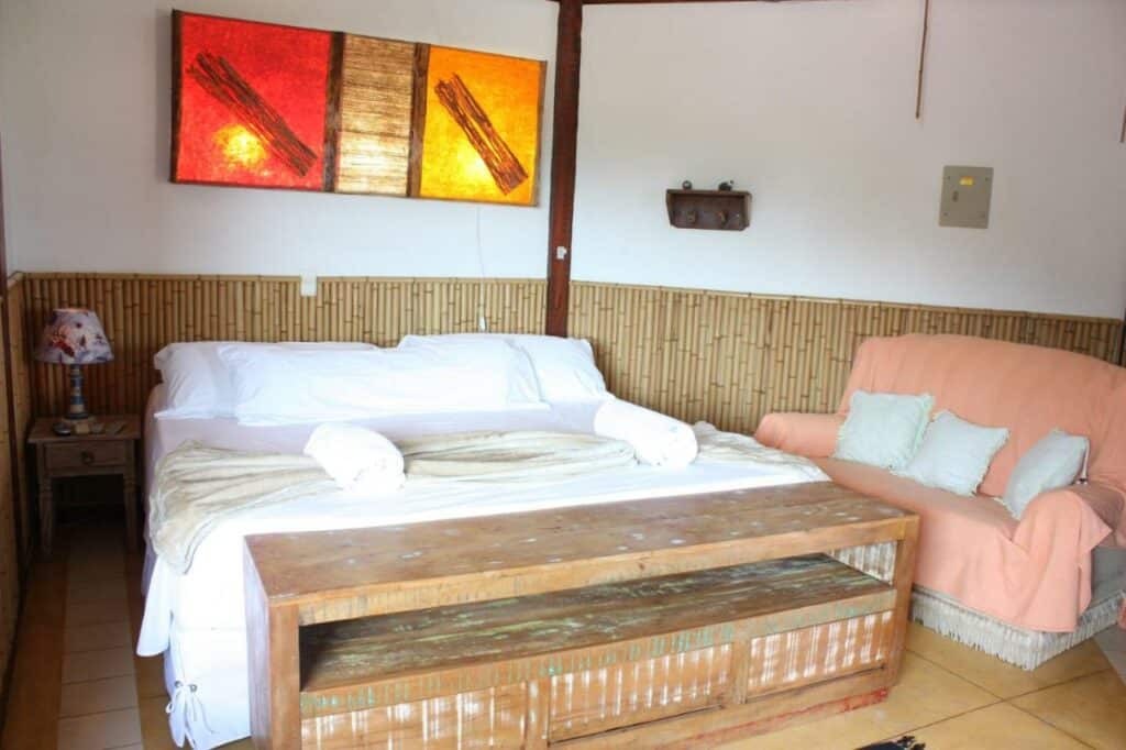 Uma cama bem arrumada com lençóis brancos, toalhas, um banco de madeira e uma poltrona coral em um quarto com paredes de bambu, da Pousada Villa Nina, em Ilhabela,