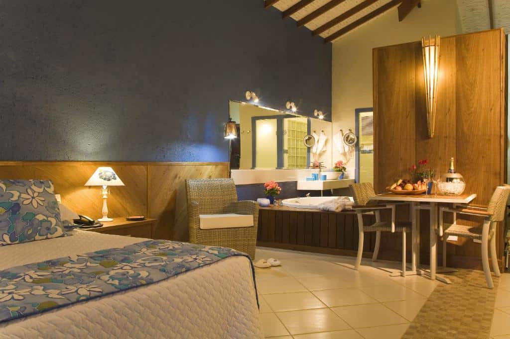 Quarto amplo da Pousada Singuitta, em parte mais afastada do centro de Ilhabela, com cama de casal, abajur e telefone na cabeceira, além de mesa com cesta de frutas e banheira de hidro