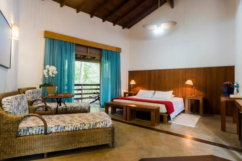 Quarto super espaçoso, com cama king size, varanda com porta de vidro e cortinas azuis semi-abertas, mostrando árvores verdes no lado de fora, e divãs, na Guest House Ilha Splendor, uma das alternativas de pousadas em Ilhabela