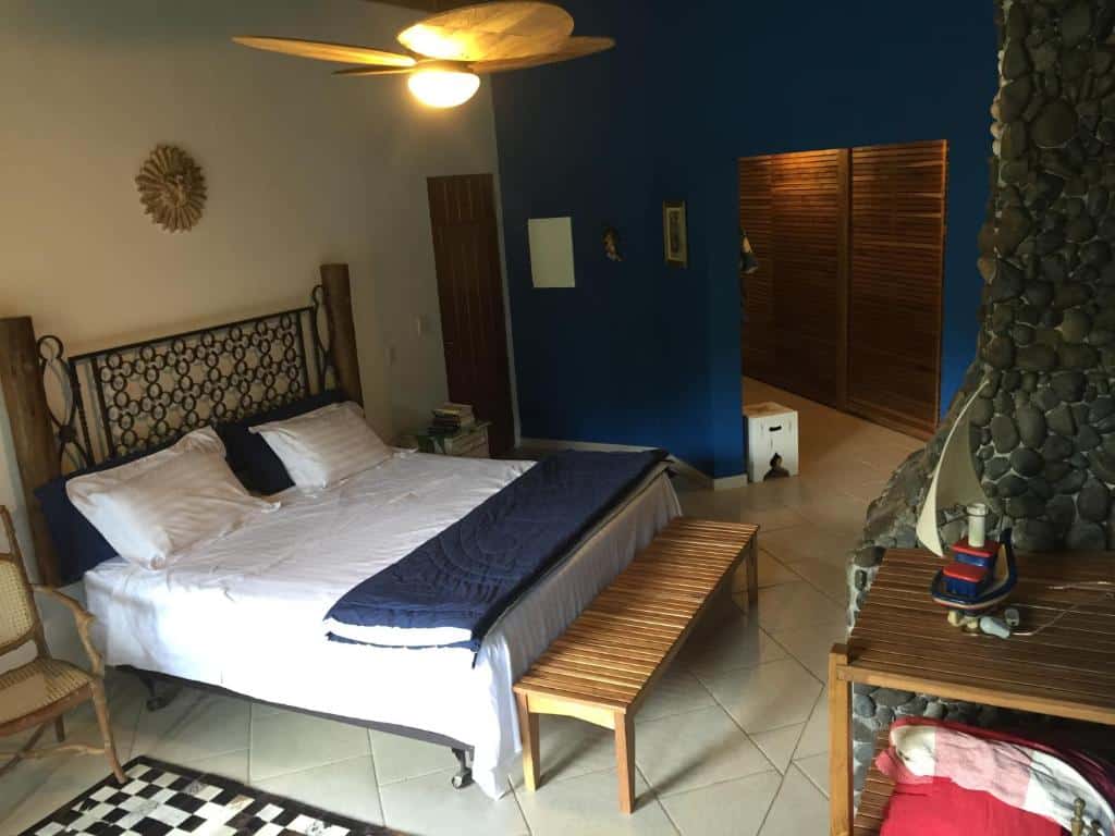Quarto da Villa Cachoeira da Rocha, uma das pousadas em Ilhabela, com cama de casal, parede com detalhes em azul, lareira de pedra, ventilador de teto e móveis de madeira.
