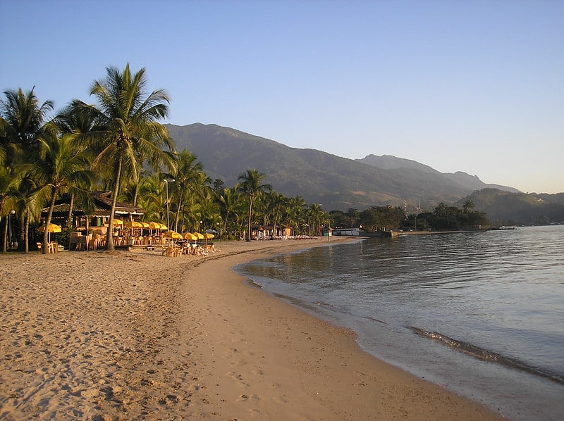 Uma praia de areia com palmeiras, guarda-sóis amarelos e uma cadeia de montanhas ao fundo sob um céu limpo.