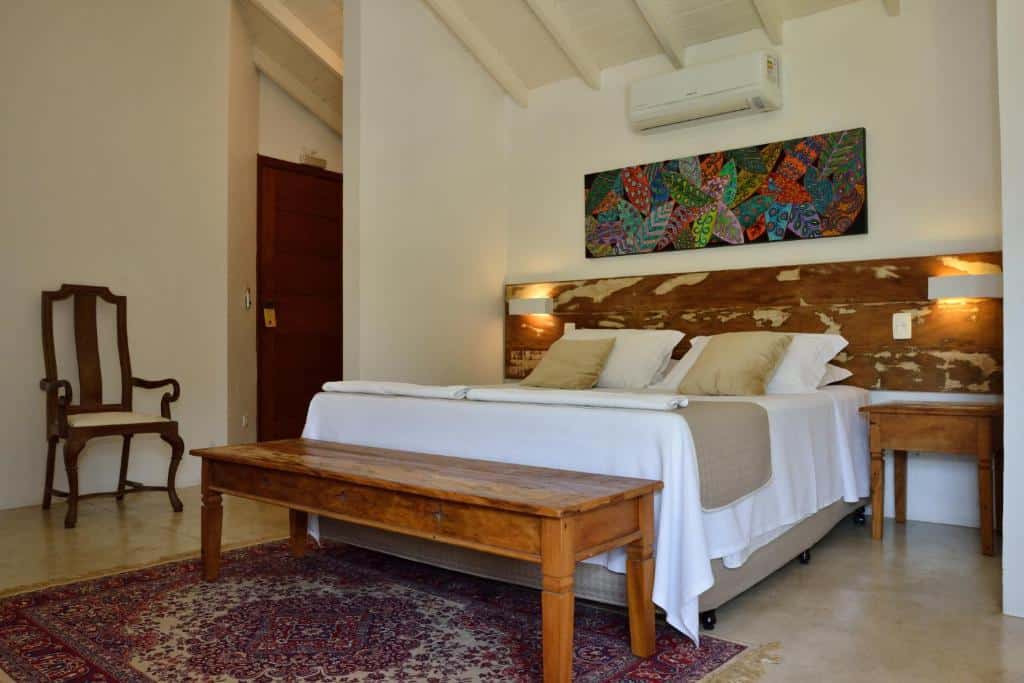 Quarto com uma cama de casal da Pousada Forte Rocha, em Ilhabela, banco de madeira, mesas laterais, uma cadeira decorativa e obras de arte coloridas acima da cabeceira da cama.