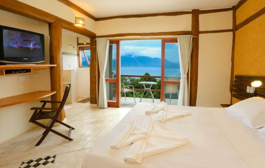 Quarto da Pousada Mariola, em Ilhabela, com uma cama de casal, TV, cadeira de madeira e varanda com vista para um lago e montanhas.