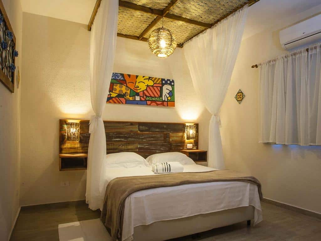 Quarto da Pousada de Charme Emirados, em Ilhabela, com cama de casal, mosqueteira, quadro colorido decorativo e luzes amarelas de cabeceira
