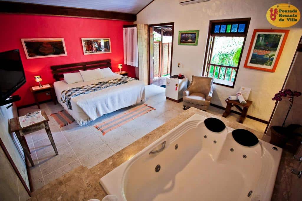 Quarto da Velinn Pousada Recanto da Vila, em Ilhabela com uma cama de casal, poltrona, banheira de hidromassagem, arte na parede e uma varanda com vegetação externa.
