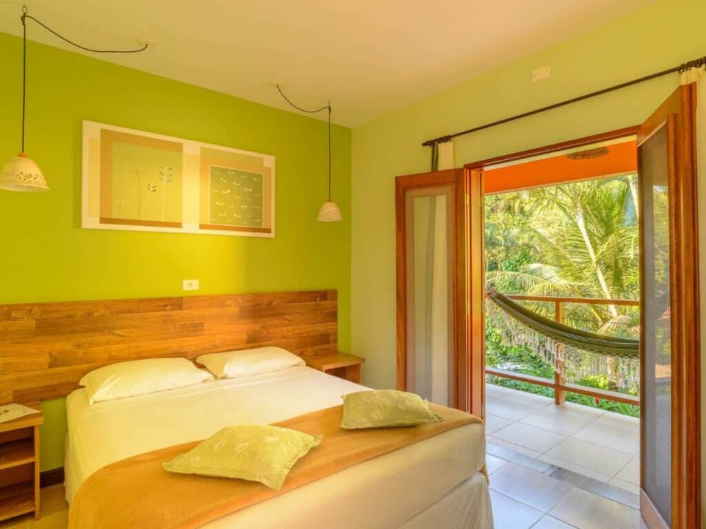 Quarto da Pousada Ecoilha, em Ilhabela, com cama de casal, parede com detalhes em verde e portas abertas que levam a uma varanda com rede e palmeiras.