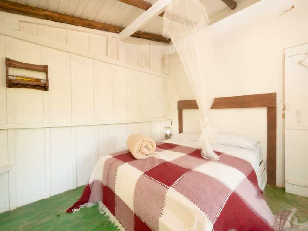 Uma cama com um cobertor xadrez vermelho e branco, duas toalhas enroladas e um mosquiteiro em um quarto com paredes brancas e piso verde, da Pousada Canto Bravo, na Praia do Bonete