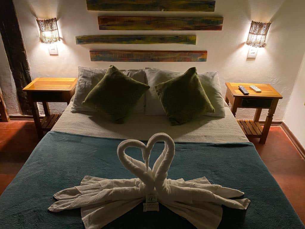 Uma cama bem arrumada da pousada Vila Pequeá Ilhabela com dois travesseiros verdes, cisnes de toalha dobrados e duas mesinhas de cabeceira de madeira com lâmpadas e controles remotos.