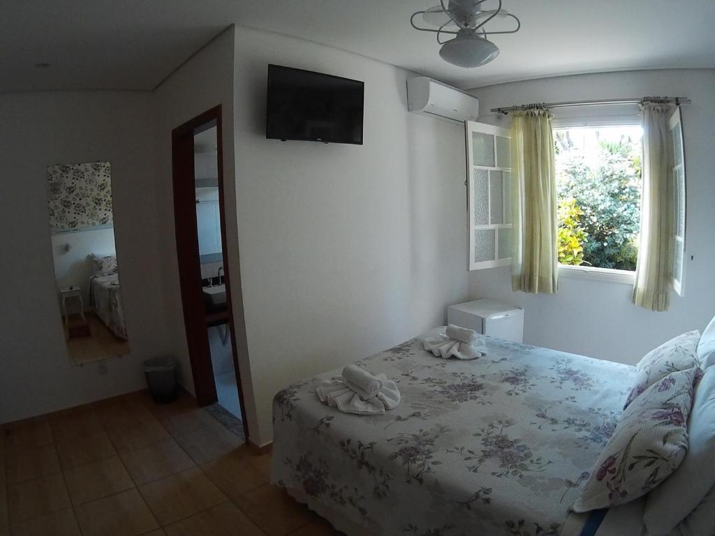 Quarto da Pousada Della Vegas com uma cama de casal, uma janela com vista para o jardim, um frigobar, uma televisão, um espelho e um banheiro, para representar pousadas em Serra Negra.