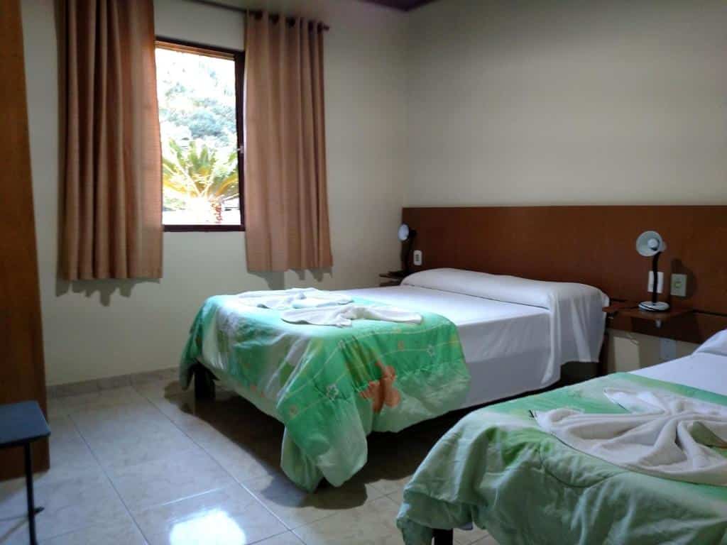 Quarto da Pousada Paraiso com uma cama de casal e uma de solteiro, uma janela com cortinas, duas mesinhas de cabeceira com luminárias.