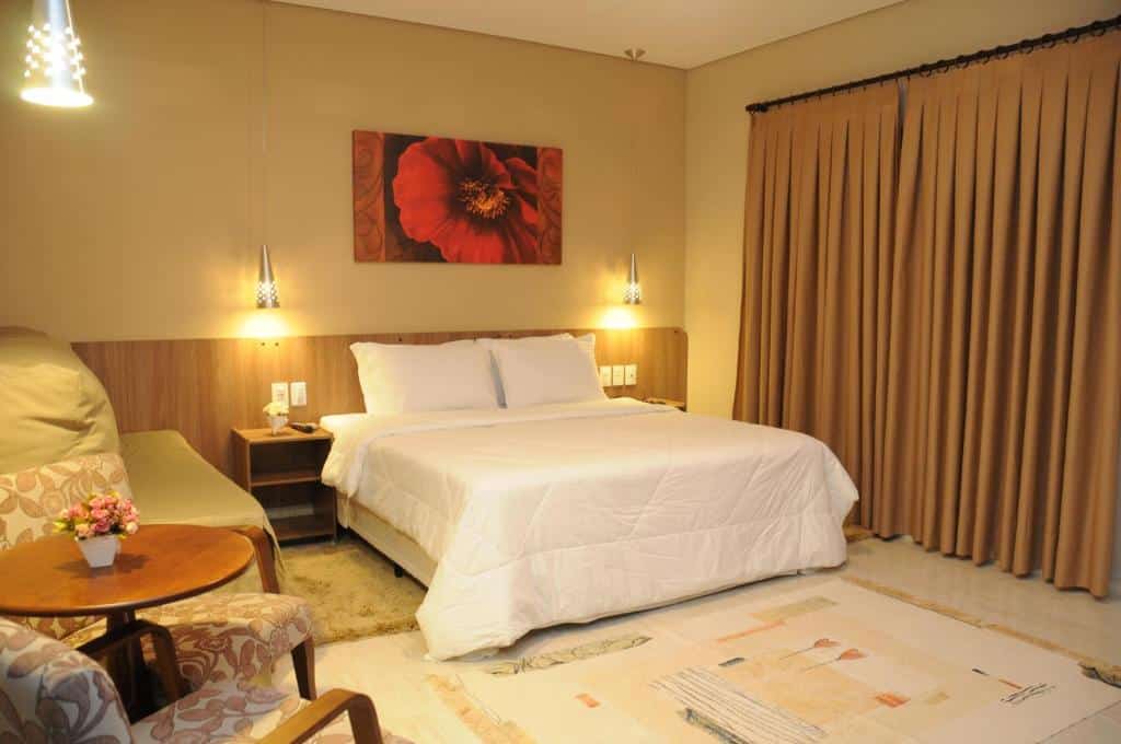 Quarto novo no O Recanto Pousada com uma cama de casa, um sofá, uma mesinha com duas poltronas, uma varanda com cortinas, tudo decorado em tons de branco, bege e madeira, para representar pousadas em Serra Negra.