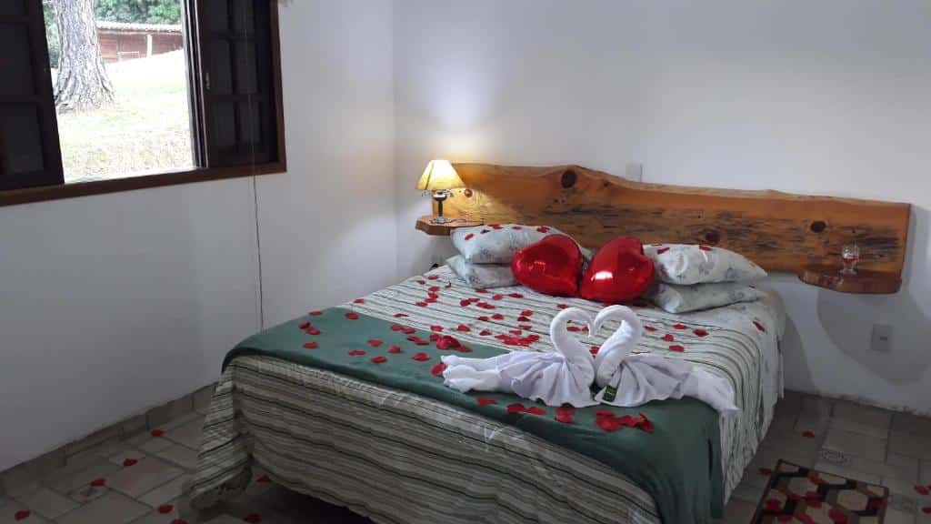 Quarto em Refúgio Serra Negra com uma cama de casal com toalhas, travesseiros e pétalas de rosas sob ela, uma cabeceira de madeira rústica com um abajur e uma janela com vista para o jardim.