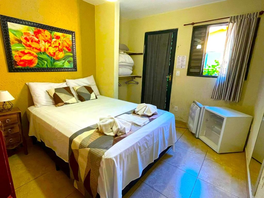 Uma cama bem arrumada em um quarto pequeno com quadro, armário, frigobar pequeno, janela e piso de azulejo, na Pousada Villa Três, para representar pousadas em Serra Negra.