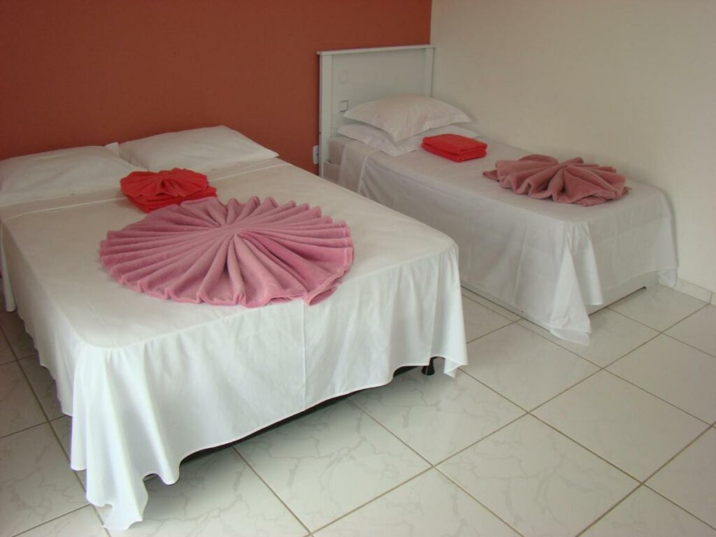 Quarto na Pousada Primavera da Serra com uma cama de casal e uma de solteiro, um terraço, tudo em tons de branco e um rosa claro.