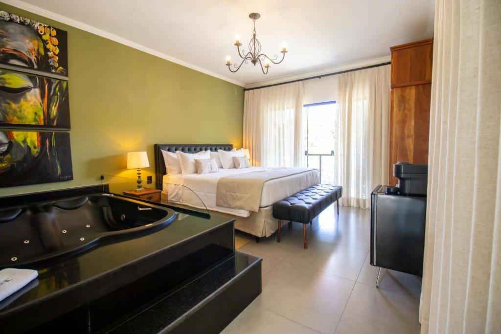 Um quarto moderno com uma cama grande, banheira de hidromassagem, banco preto, obras de arte, parede verde e cortinas transparentes cobrindo uma janela, na Unalome Pousada, para representar pousadas em Serra Negra.