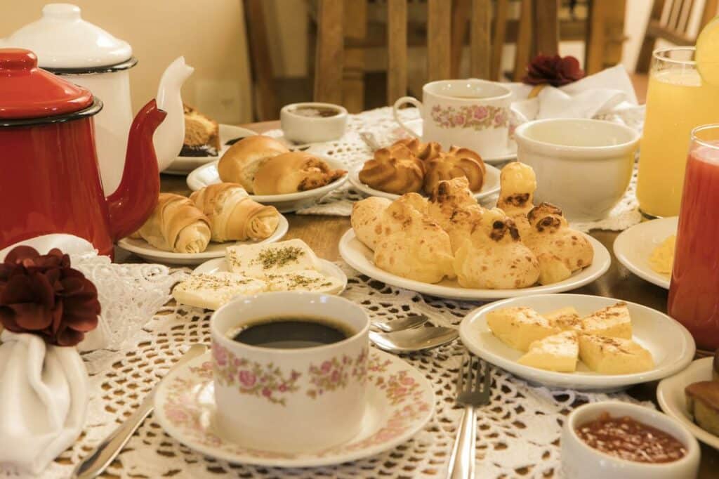 Mesa posta da Pousada Encanto da Serra com café, suco, pão, bolos, queijo e geleia em jogos americanos de renda e pratos com estampas florais.