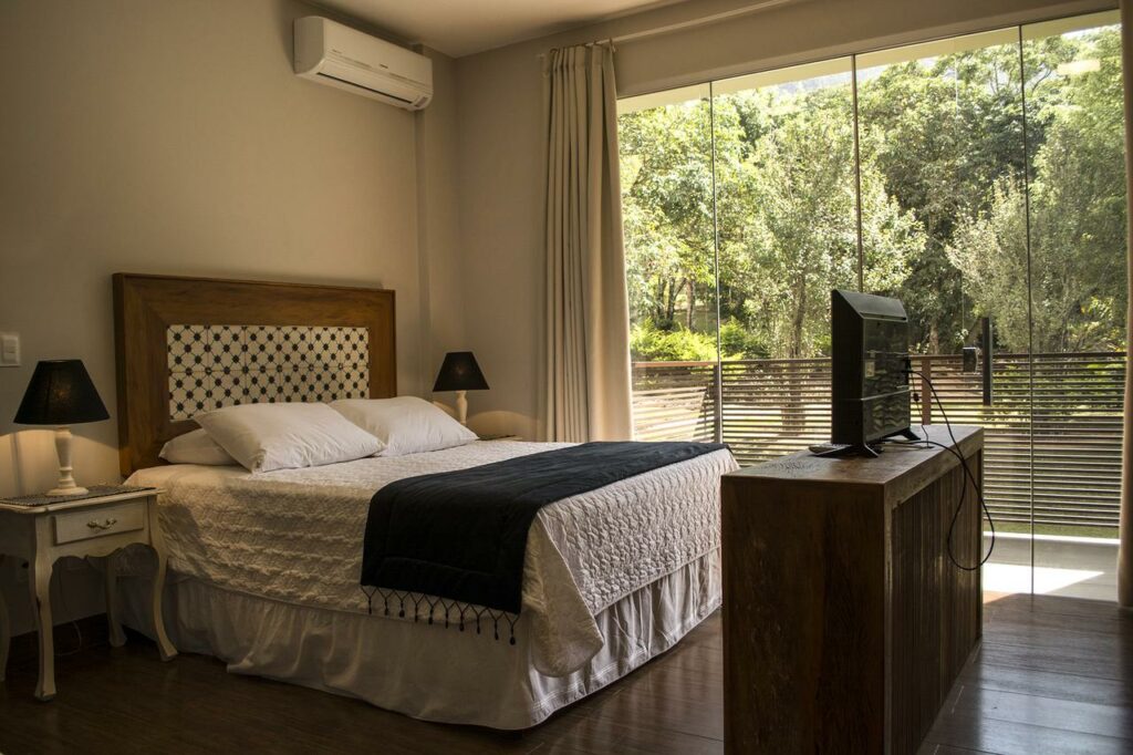 Quarto da Pousada Recanto das Araras, uma das pousadas em Tiradentes, com uma cama bem arrumada, TV em um suporte de madeira e uma grande janela com vista para árvores e vegetação.