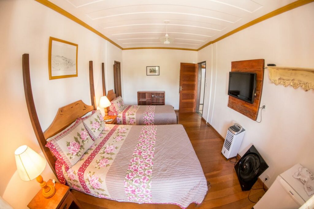 Quarto da Pouso Lar Doce Lar, uma das pousadas em Tiradentes, com duas camas de casal, roupa de cama floral, uma TV na parede, luminárias e piso de madeira.