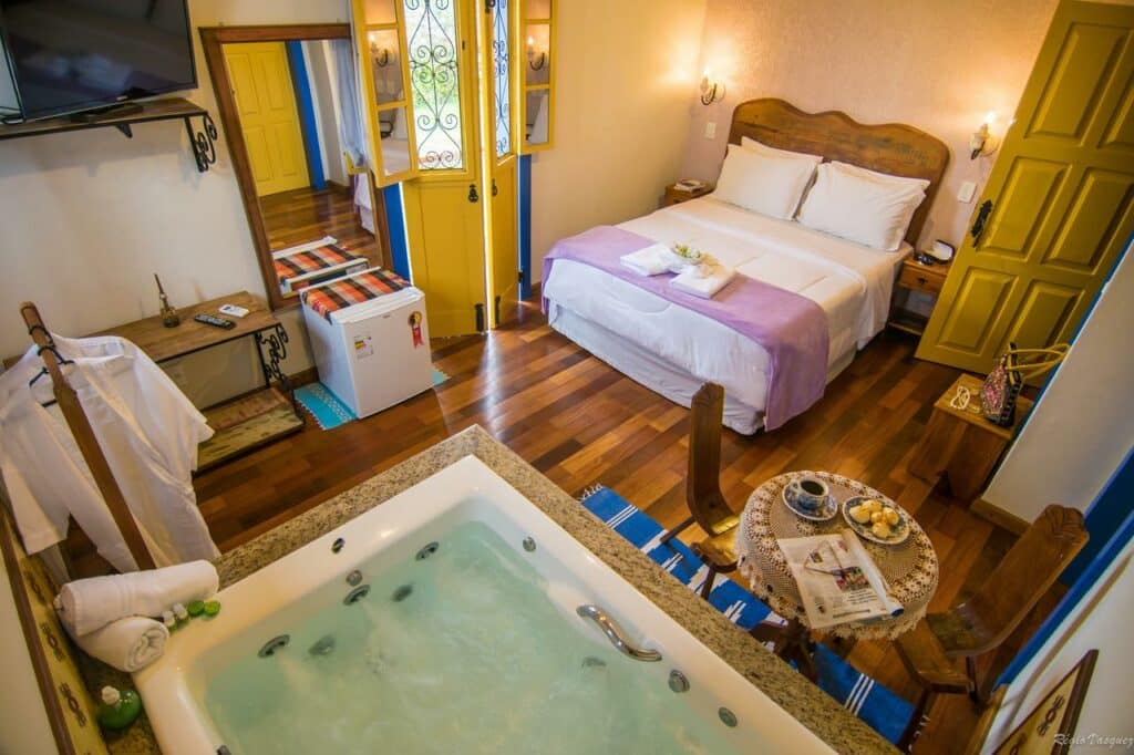 Quarto da Pousada Marília de Dirceu, uma das pousadas em Tiradentes, com uma cama de casal, uma pequena mesa com cadeiras, uma banheira de hidromassagem e uma TV, com piso de madeira e portas amarelas.