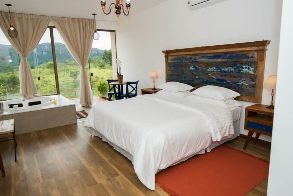 Quarto da pousada Solar Da Serra Tiradentes com uma cama grande, piso de madeira, banheira, mesas de cabeceira, luminárias e janelas grandes com vista para a vegetação.