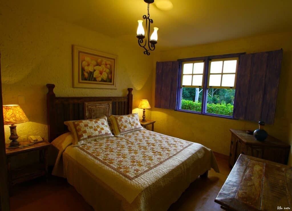 Quarto aconchegante de A Casa Vermelha Hospedaria, uma das pousadas em Tiradentes, com uma cama de madeira, duas luminárias, uma colcha acolchoada e uma janela com persianas azuis que mostram a vegetação do lado de fora.
