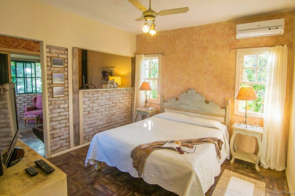 Quarto iluminado da Pousada Villa Allegra pelo sol com uma cama de casal, duas mesas de cabeceira, luminárias, um ventilador de teto e uma parede divisória de tijolos expostos.