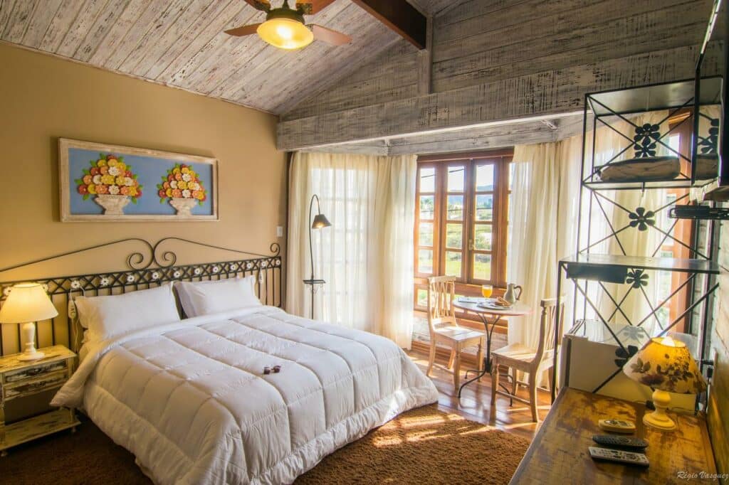 Quarto aconchegante da Pousada Trem do Imperador, em Tiradentes, com uma cama de casal, teto de madeira, pequena mesa de jantar perto de uma janela e iluminação natural aconchegante.