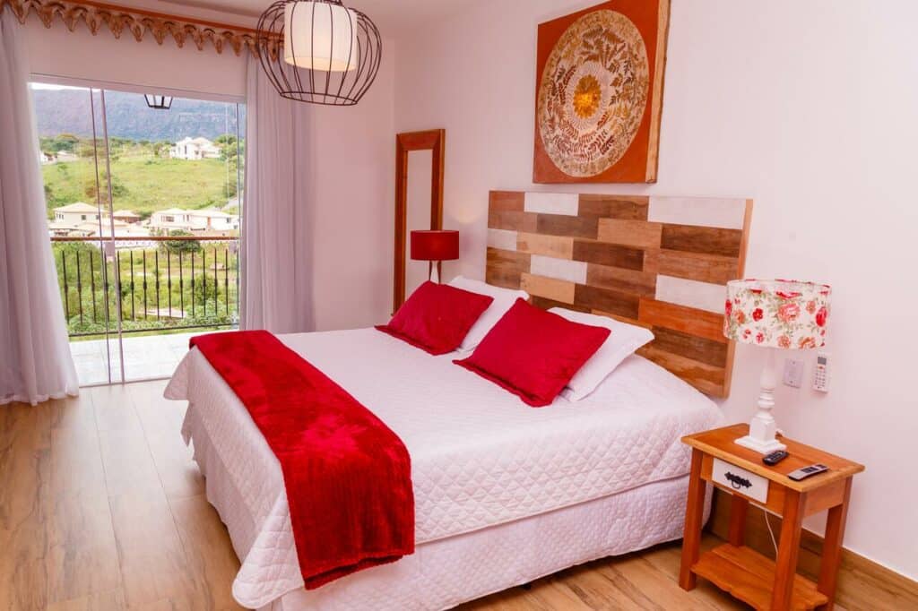 Quarto com cama de casal da Chalés Pôr do Sol, uma das pousadas em Tiradentes, com detalhes em vermelho, cabeceira de madeira, mesa de cabeceira, luminária, arte na parede e varanda com vista.