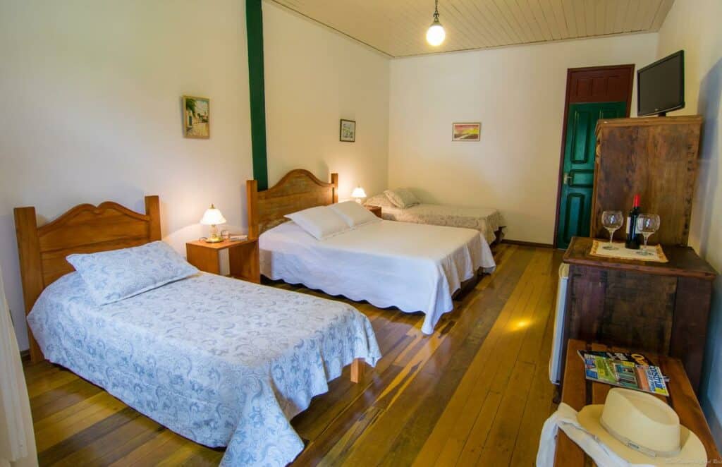 quarto da Pousada da Trilha, uma das pousadas em Tiradentes, com uma cama de solteiro e duas camas de casal, piso de madeira, mesas de cabeceira, arte na parede, TV e uma cômoda com itens.